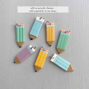 May include: A collection of colorful pencil charms in various pastel shades, including blue, yellow, green, and purple. Each charm features a black tip, a pink eraser, and a silver keyring. The text on the image reads: "add on pencil charms ~ sold separately in my shop:)"