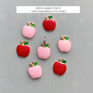 May include: A collection of apple charms in red and pink with gold-colored stems and green leaves. The charms are arranged on a light gray surface. The text in the image says "add an apple charm - sold separately in my shop:)"