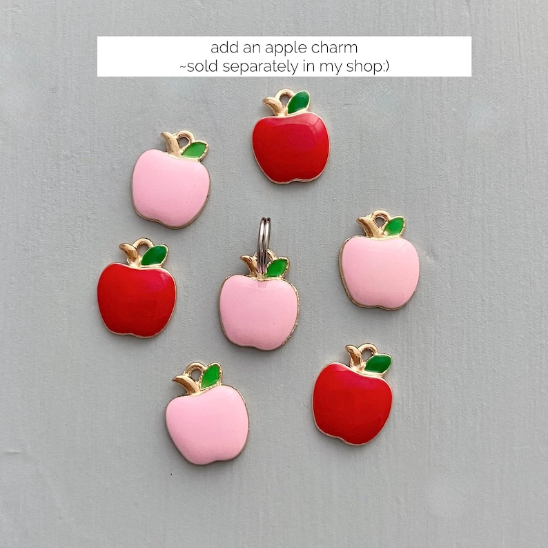 May include: A collection of apple charms in red and pink with gold-colored stems and green leaves. The charms are arranged on a light gray surface. The text in the image says "add an apple charm - sold separately in my shop:)"