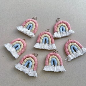 May include: Six macrame rainbow ornaments featuring three concentric arches in pastel pink, cream, and light blue cotton cord. Each rainbow includes a silver metal jump ring at the top and a frayed white fringe at the bottom edge of the arches.