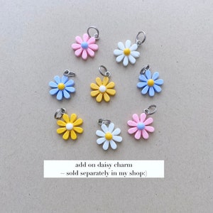 May include: A collection of daisy flower charms in various colors, including pink, white, yellow, and blue, each with a silver-colored loop. The charms have a pastel color palette and are arranged on a neutral background. Text on the image reads: "add on daisy charm ~ sold separately in my shop:)"