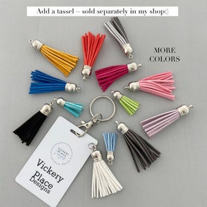 May include: A collection of colorful suede tassels with silver-tone caps, key rings, and a white tag with the text "Vickery Place Designs". Tassels are in various colors, including yellow, red, blue, pink, and black. The text "MORE COLORS" is also visible.