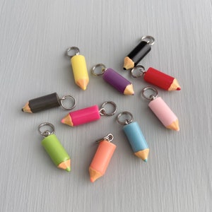 May include: A collection of colorful pencil-shaped keychains in various colors, including yellow, black, red, pink, green, and blue. Each keychain has a silver ring and a pencil tip. The keychains are arranged on a light gray surface.