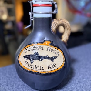 May include: A dark gray ceramic beer growler with a swing top closure. The label reads "Dogfish Head Punkin Ale" and features a shark illustration. The handle is made of a woven material.