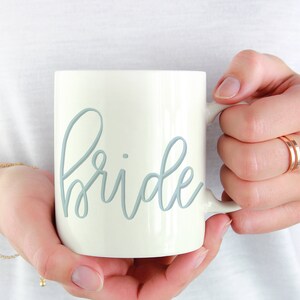May include: White ceramic mug with a light blue cursive inscription that reads "bride".