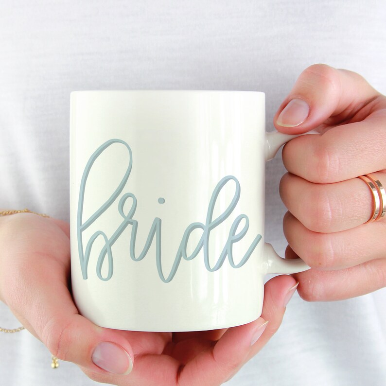 May include: White ceramic mug with a light blue cursive inscription that reads "bride".