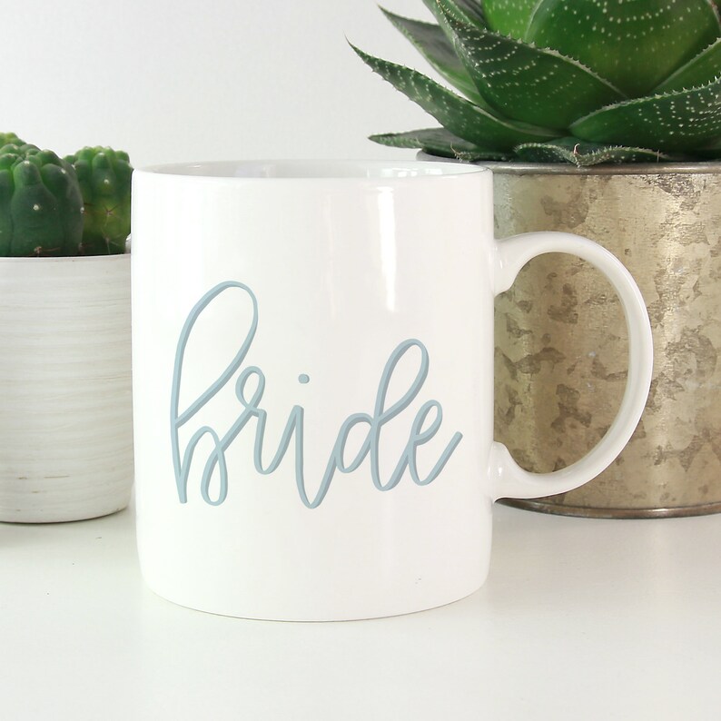 May include: White ceramic mug with a blue cursive inscription that reads "bride".