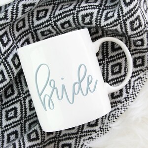 May include: White ceramic mug with a blue cursive inscription that reads "bride". The mug is sitting on a black and white patterned blanket.