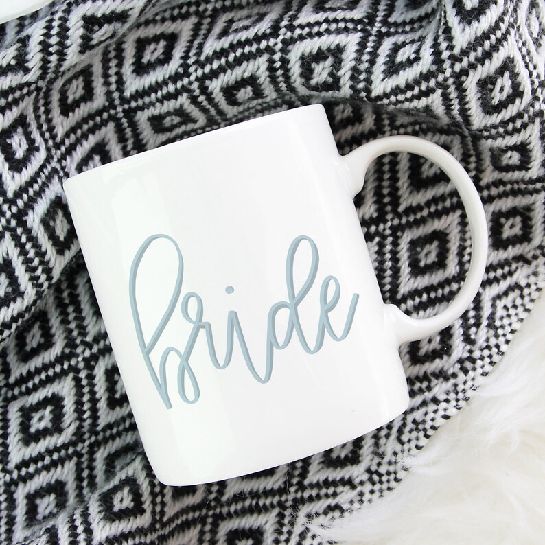 May include: White ceramic mug with a blue cursive inscription that reads "bride". The mug is sitting on a black and white patterned blanket.