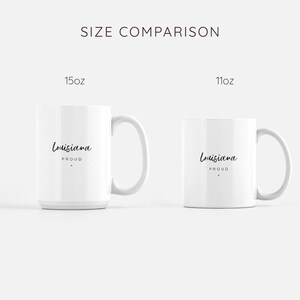 May include: Size comparison of two white ceramic mugs. The larger mug is 15 oz and the smaller mug is 11 oz. Both mugs feature black text that reads "Louisiana Proud".