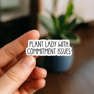 May include: A white sticker with the text "PLANT LADY WITH COMMITMENT ISSUES" in black, held in a hand. The sticker is rectangular with rounded corners. The background is blurred, featuring a green plant in a blue pot.