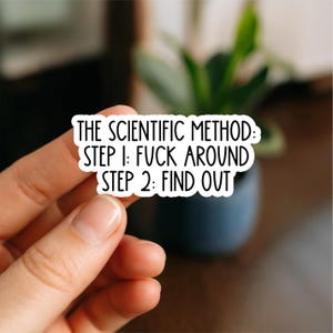 May include: A white sticker with black text that reads "THE SCIENTIFIC METHOD: STEP 1: FUCK AROUND STEP 2: FIND OUT." The sticker is held by a hand in front of a blurred background with a plant.