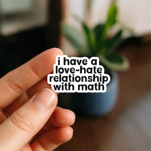 May include: A white sticker with a black border and the text "i have a love-hate relationship with math". The sticker is held in a person's hand, with a blurred background of a plant in a dark pot.