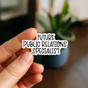 May include: A white sticker with black text that reads "FUTURE PUBLIC RELATIONS SPECIALIST." The sticker is held between fingers, with a blurred background of a plant in a dark blue pot.