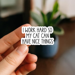May include: A white sticker with black text that reads "I WORK HARD SO MY CAT CAN HAVE NICE THINGS." The sticker is held between fingers, with a blurred background of a plant in a dark pot.