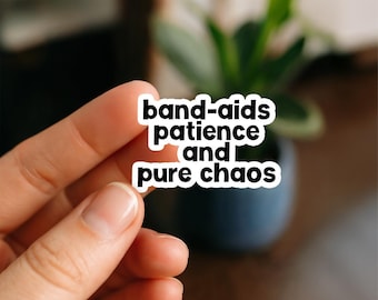 May include: A white sticker with black text that reads "band-aids patience and pure chaos." The sticker is held between two fingers. A blurred background includes a plant in a dark blue pot.