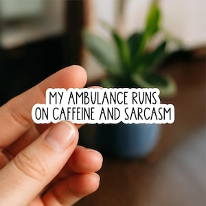May include: A white sticker with black text that reads "MY AMBULANCE RUNS ON CAFFEINE AND SARCASM." The sticker is held by a hand, with a blurred background of a plant and a dark blue pot.