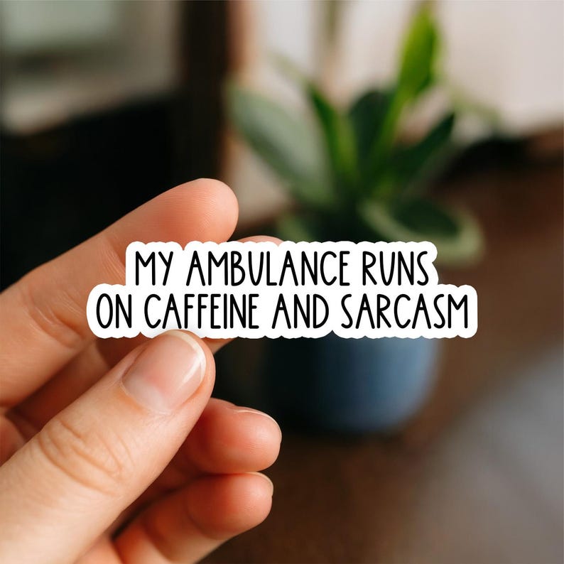 May include: A white sticker with black text that reads "MY AMBULANCE RUNS ON CAFFEINE AND SARCASM." The sticker is held by a hand, with a blurred background of a plant and a dark blue pot.