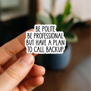 May include: A white sticker with black text that reads "BE POLITE BE PROFESSIONAL BUT HAVE A PLAN TO CALL BACKUP". The sticker is held by a hand in front of a blurred background.
