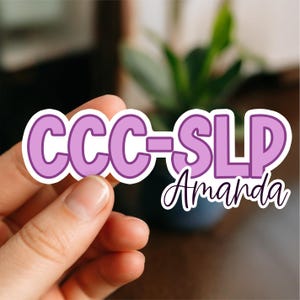 May include: A sticker featuring the text "CCC-SLP Amanda" in purple and white. The sticker is held by a hand. The letters "CCC-SLP" are in a bold, sans-serif font, while "Amanda" is in a cursive script.