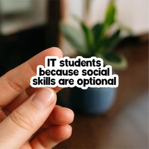 Peut inclure: Un autocollant blanc avec du texte noir qui dit "IT students because social skills are optional". L'autocollant est tenu par une main, avec un arrière-plan flou d'une plante et d'un pot bleu foncé.