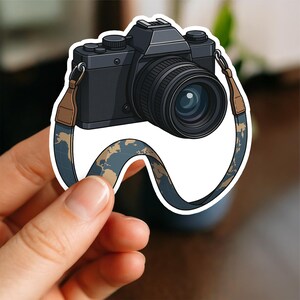 May include: A black camera sticker with a lens and a strap featuring a world map design. The strap is brown and blue. The camera is held by a hand against a white background.