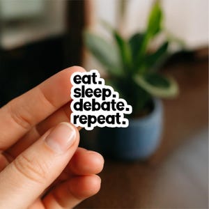 May include: A white sticker featuring the text "eat. sleep. debate. repeat." in a black, stacked font. The sticker is held by a hand, with a blurred background of a plant in a dark blue pot.