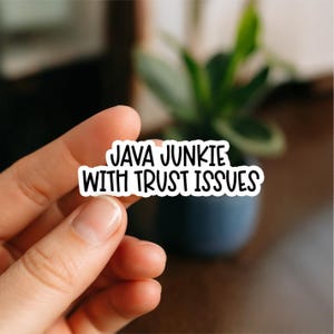 Può includere: Un adesivo bianco con testo nero che recita "JAVA JUNKIE WITH TRUST ISSUES". L'adesivo è tenuto in mano, con uno sfondo sfocato di una pianta in un vaso blu scuro.