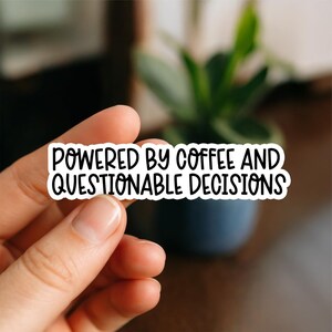 May include: A white sticker with black text saying "POWERED BY COFFEE AND QUESTIONABLE DECISIONS." The sticker is held between fingers, with a blurred background of a plant in a blue pot.