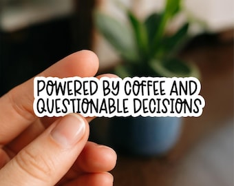 May include: A white sticker with black text that reads "POWERED BY COFFEE AND QUESTIONABLE DECISIONS." The sticker is held between fingers, with a blurred background of a plant in a blue pot.