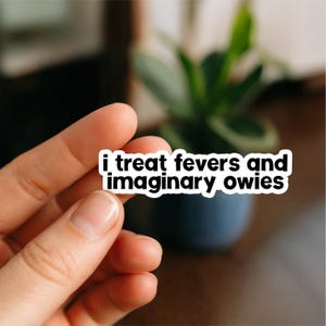 May include: A white sticker with black text that reads "i treat fevers and imaginary owies." The sticker is held in a person's hand, with a blurred background of a plant in a blue pot.