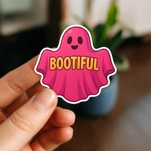 May include: A pink ghost sticker with the word "BOOTIFUL" in yellow letters. The ghost has a smiling face and a white border. The sticker is a fun, festive item, perfect for Halloween.