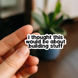 Peut inclure: Un autocollant blanc avec du texte noir qui dit "i thought this would be about building stuff." L'autocollant est tenu par une main, avec un arrière-plan flou d'une plante dans un pot bleu foncé.