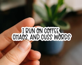 May include: A white sticker with black text that reads "I RUN ON COFFEE, CHAOS, AND CUSS WORDS." The sticker is held by a hand in front of a blurred background with a plant.