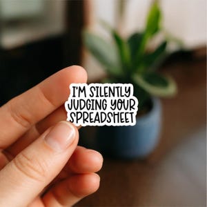 May include: A white sticker with black text that reads "I'M SILENTLY JUDGING YOUR SPREADSHEET." The sticker is held between fingers, with a blurred background of a plant in a dark blue pot.