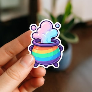May include: A colorful sticker featuring a cartoon cauldron with a rainbow design and purple accents. The cauldron has a pink and blue vapor rising from it. The sticker is held by a hand.