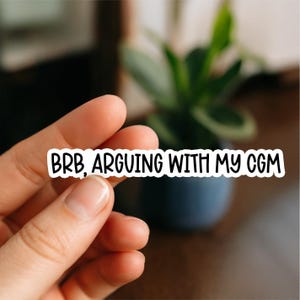 May include: A white sticker with black text that reads "BRB, ARGUING WITH MY CGM." The sticker is held between fingers, with a blurred background of a plant in a dark blue pot.