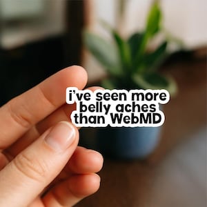 May include: A white sticker with black text that reads "I've seen more belly aches than WebMD." The sticker is held by a hand, with a blurred background of a plant and a dark blue pot.
