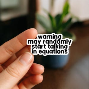 May include: A white sticker with black text that reads "warning may randomly start talking in equations." The sticker is held in front of a blurred background of a plant in a dark blue pot.