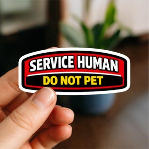 May include: A rectangular sticker with a white border, red background, and black accents. The text "SERVICE HUMAN" is in white, above "DO NOT PET" in yellow. The sticker is held by a hand.