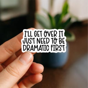 May include: A white sticker with black text that reads "I'll get over it just need to be dramatic first." The sticker is held by a hand, with a blurred background of a plant and a dark surface.