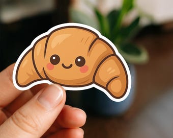 May include: A sticker of a cartoon croissant with a smiling face. The croissant is light brown with darker brown details and pink cheeks. The sticker has a white border.