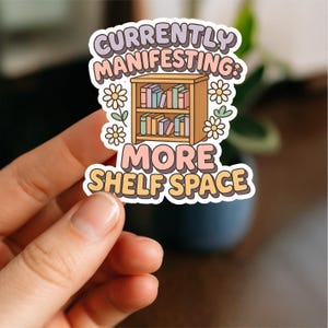 May include: A sticker with the text "Currently Manifesting: More Shelf Space" in a retro font. The sticker features a cartoon bookshelf with books and daisy flowers. The sticker is held by a hand.