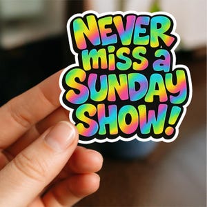 May include: A colorful sticker with the text "NEVER miss a SUNDAY SHOW!" in rainbow tie-dye lettering. The sticker has a black background and a white border. The sticker is held by a person.