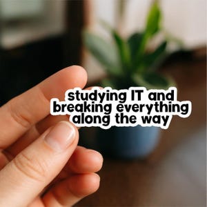 Peut inclure: Un autocollant blanc avec du texte noir qui dit "studying IT and breaking everything along the way". L'autocollant est tenu par une main, avec un arrière-plan flou d'une plante dans un pot bleu foncé.
