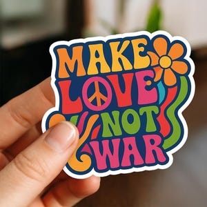 Könnte beinhalten: Ein farbenfroher Aufkleber mit dem Schriftzug "MAKE LOVE NOT WAR" in einem Retro-Schriftstil. Das Design beinhaltet ein Friedenssymbol und eine Blume mit blauem Rand. Der Aufkleber wird von einer Hand gehalten.