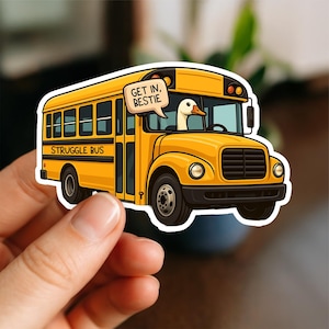 May include: A yellow school bus sticker with the words "STRUGGLE BUS" on the side. A cartoon duck is driving the bus and has a speech bubble that says "GET IN, BESTIE". The sticker is held by a hand.