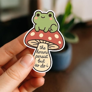 Peut inclure: Un autocollant représentant une grenouille verte de dessin animé assise sur un champignon rouge à pois blancs. La tige du champignon porte l'inscription "the horrors persist but so do i". La grenouille a des joues roses. L'autocollant est tenu par une main.