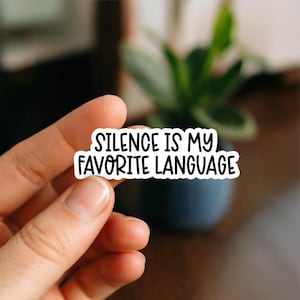 Könnte beinhalten: Ein weißer Aufkleber mit schwarzem Text, der lautet: „SILENCE IS MY FAVORITE LANGUAGE“. Der Aufkleber wird zwischen den Fingern gehalten, mit einem verschwommenen Hintergrund einer Pflanze und einem dunkelblauen Topf.