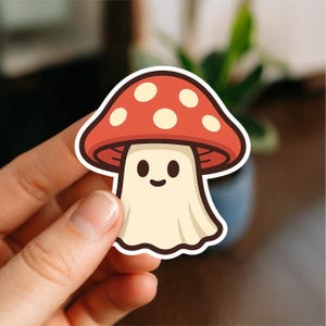 May include: A sticker featuring a cartoon mushroom with a red cap dotted with white spots. The mushroom has a smiling face and a cream-colored body. The sticker is held by a hand.